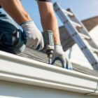 A skilled handyman wearing protective gloves is installing a new roof guttering system He is using a power drill to fasten the gutter securely to the roof